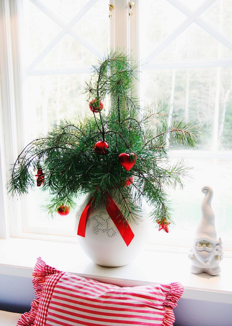 Rote Weihnachtsbaumanhänger an Kieferzweigen in Vase und Porzellan-Zwerg auf Fensterbrett