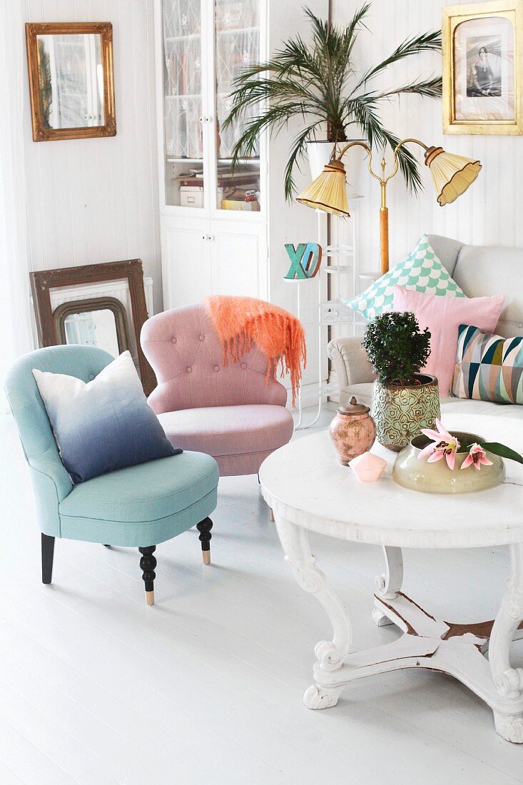 Pastel easy chairs, white, round coffee table with carved base, standard lamp and potted palm on plant stand