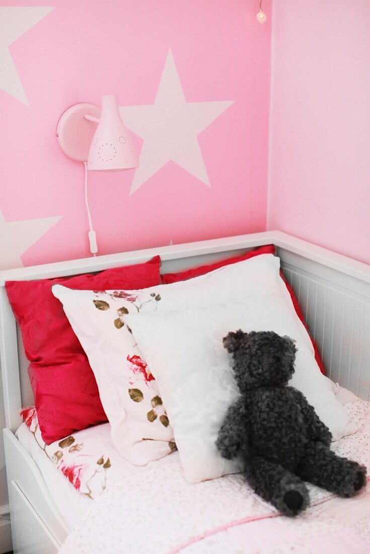 Weisses Kinderbett aus Holz mit Kissen und Teddybär, Wandleuchte auf rosa Wand mit Sternmotiv