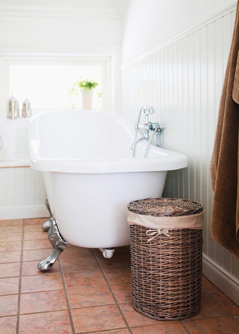Laundry basket and free-standing, vintage clawfoot bathtub against wainscoting in white bathroom