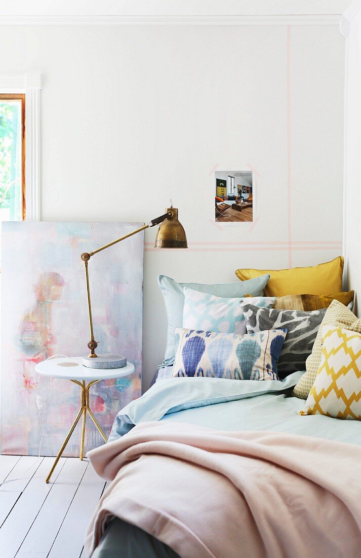 Kissenstapel auf Bett und Beistelltisch mit Messing Tischleuchte im Retro Stil vor Wand mit pastellfarbenem Streifenmuster aus Masking Tape
