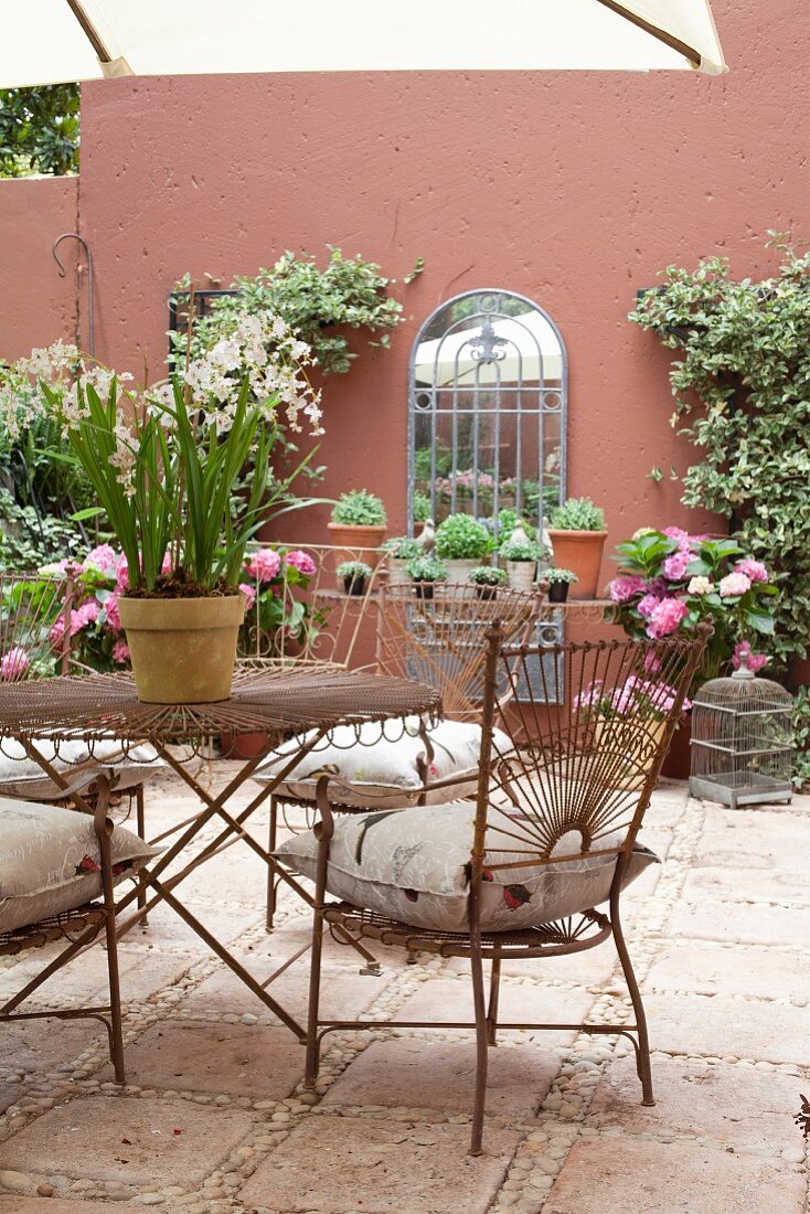 Filigrane Metallstühle und passender Tisch im Innenhof auf Fliesenboden, im Hintergrund Pflanzentöpfe vor rotbrauner Fassade