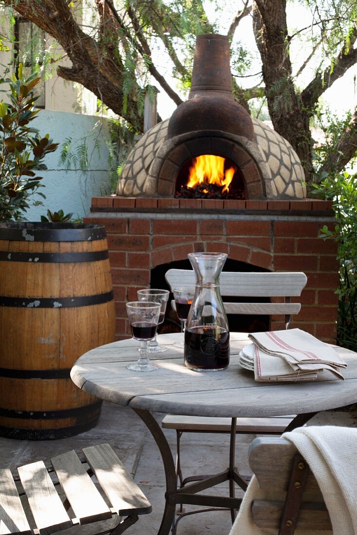Weingläser und Karaffe auf Bistrotisch vor gemauertem Pizzaofen mit loderndem Feuer