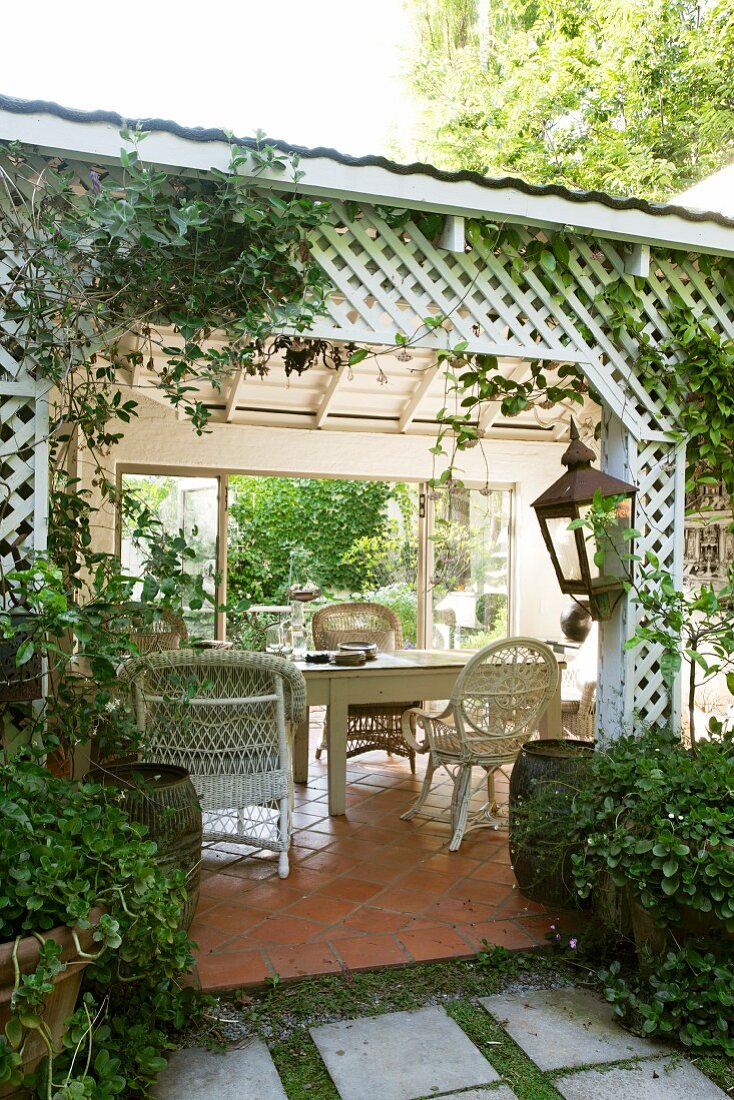 weiße Korbstühle und Holztisch auf überdachter Terrasse mit umlaufendem, kreuzförmigem Rankgitter und Terracottafliesen im Landhausstil