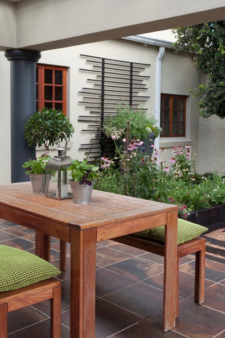 Tisch- und Bankgarnitur aus Holz, grüne Sitzpolster, auf gefliester Terrasse, im Hintergrund Anbau mit Rankhilfe auf Fassade