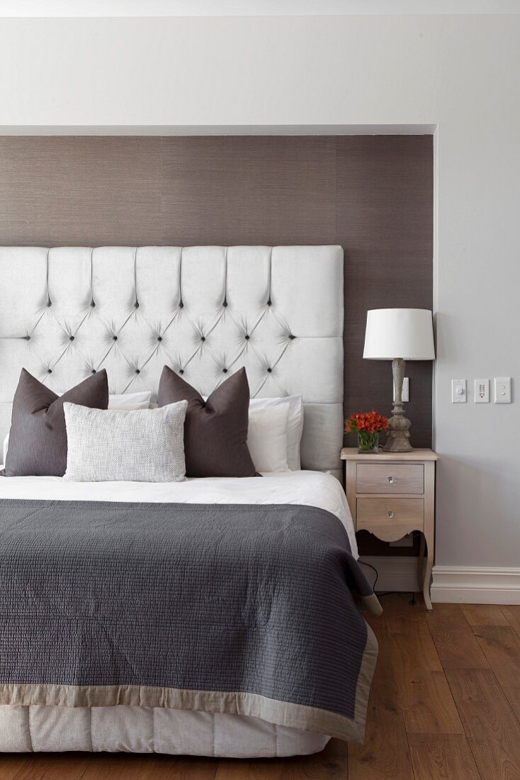 Elegant bed with scatter cushions arranged against button-tufted headboard in niche with dark brown fabric wallpaper