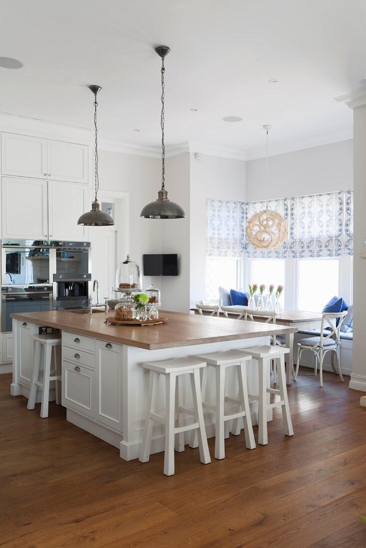 Free-standing island counter with white bar stools in open-plan country-house kitchen with dining set in window bay