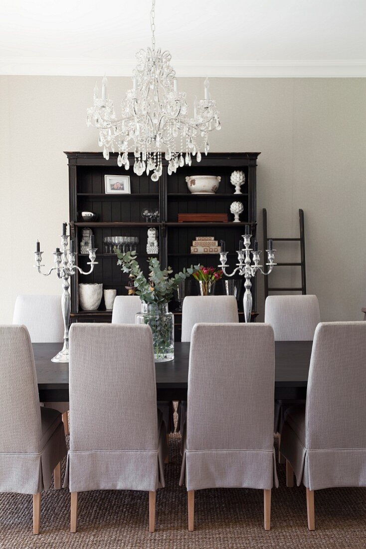 Chairs with pale grey loose covers around dining table below chandelier with glass pendants; antique, dark wood shelves in background
