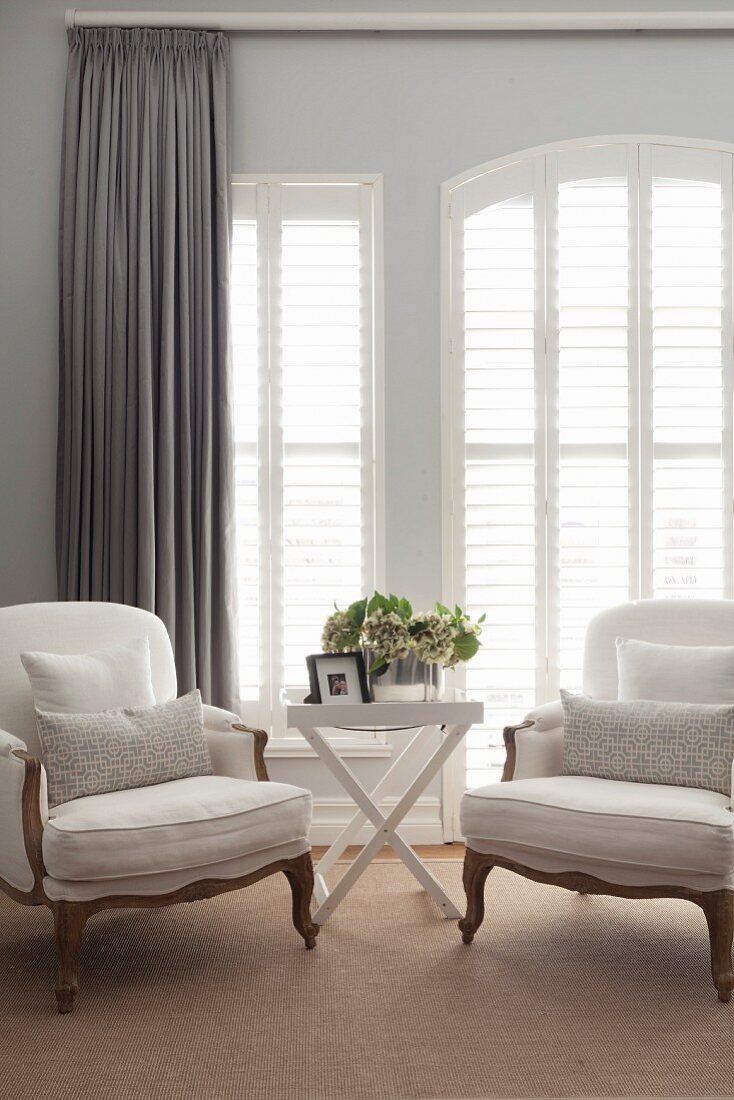 Rococo-style white armchairs and tray table in front of French windows with closed interior shutters