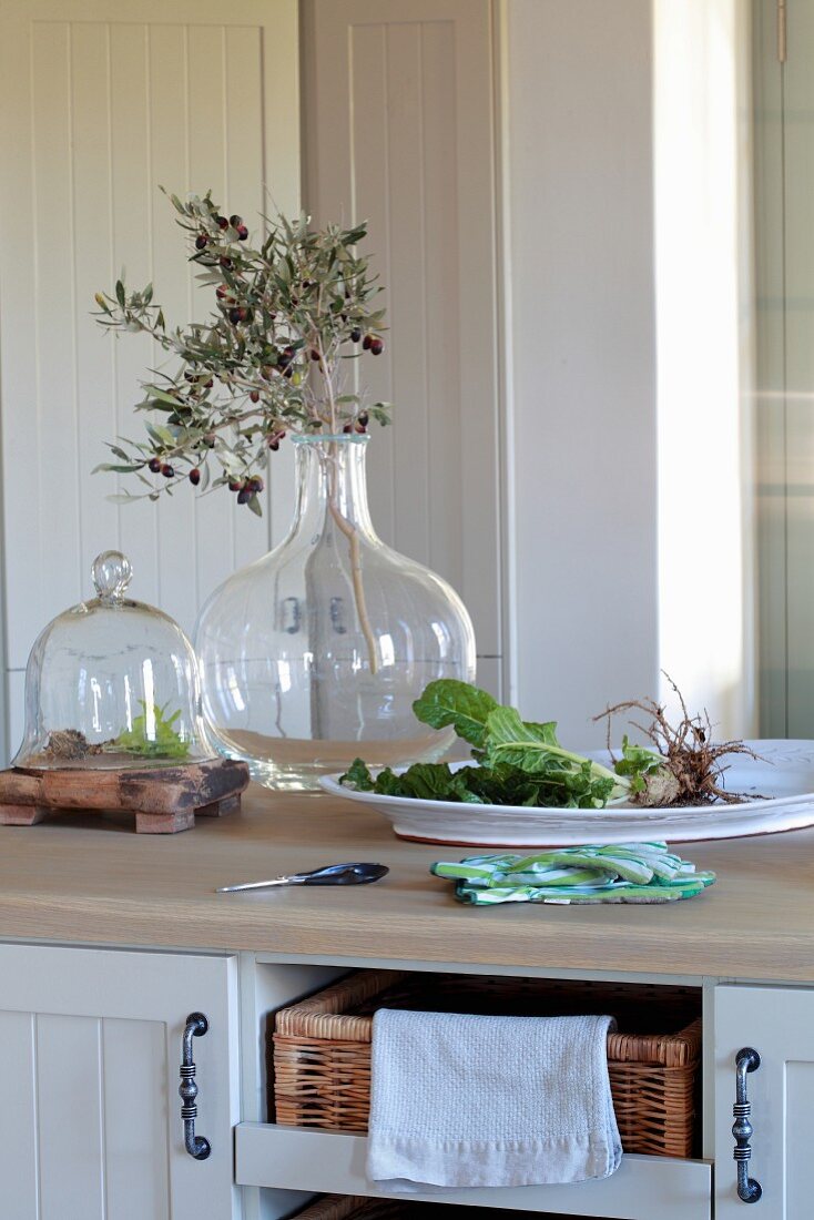 Glasvase mit Olivenzweig neben Schale mit frischem Gemüse auf Küchentheke