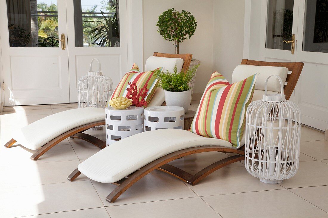 Zwei Liegestühle aus Holz mit bunt gestreiften Kissen auf einer Veranda, Laternen und Koralle als Deko