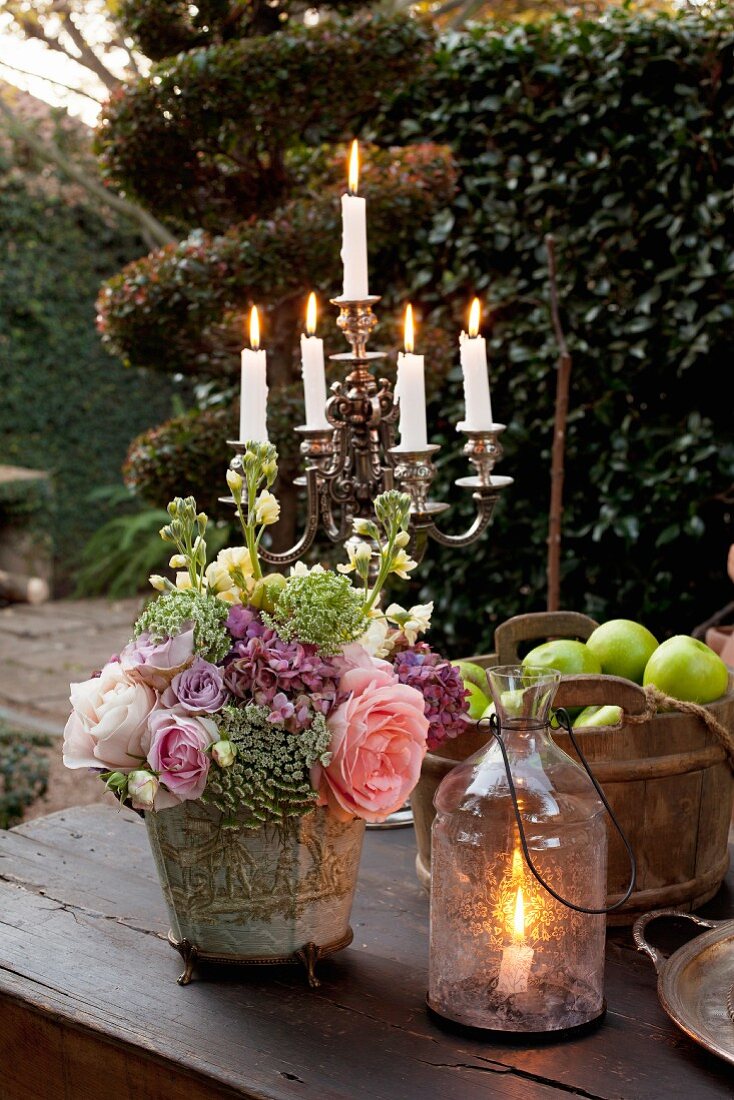 Windlicht und Blumengesteck vor mehrarmigen Kerzenleuchter auf rustikalem Gartentisch