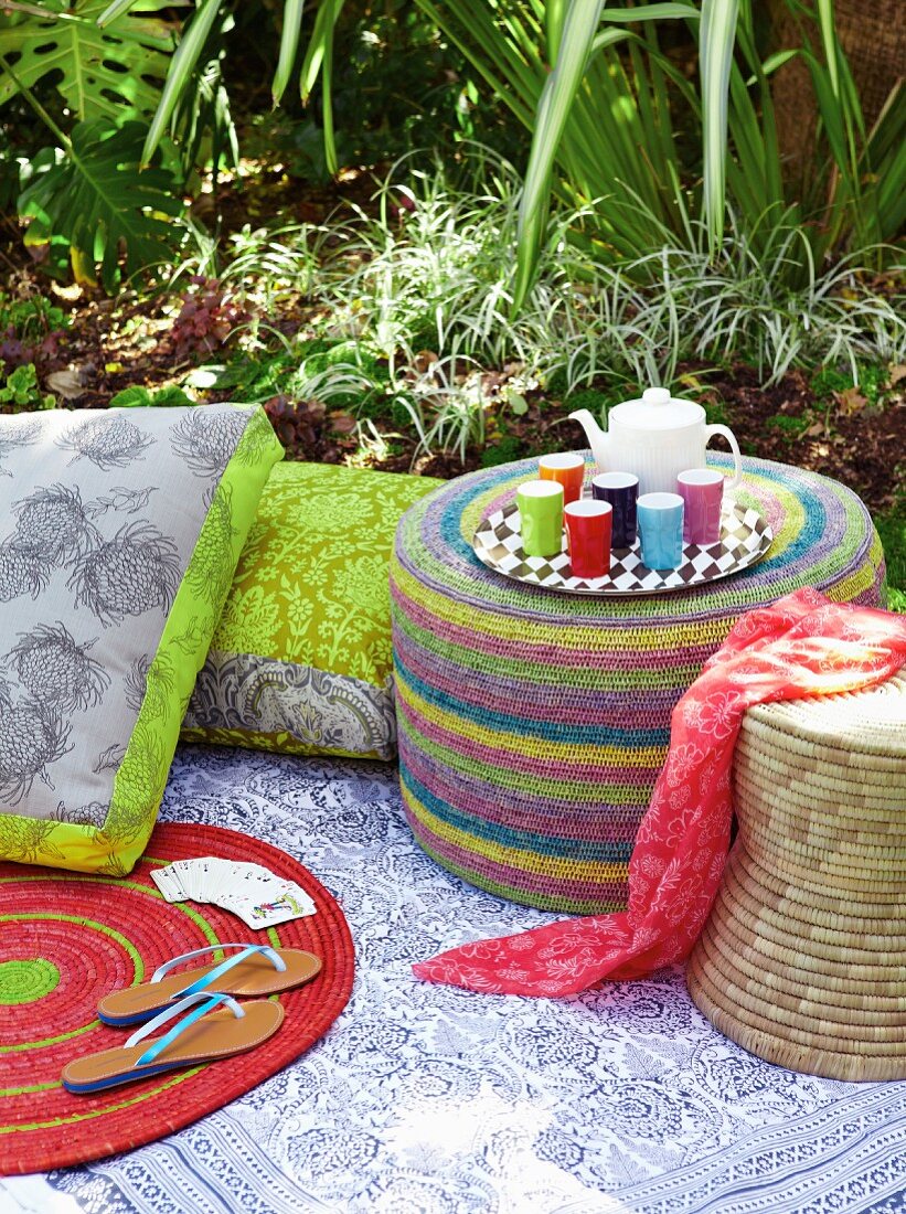Sitzpouf, Flechthocker und floralen Sitzkissen auf Picknickdecke im Garten