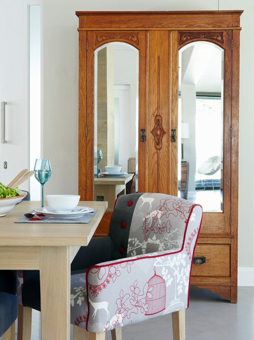 Armchair with modern upholstery at dining table with place setting; antique wardrobe with mirrored doors in background