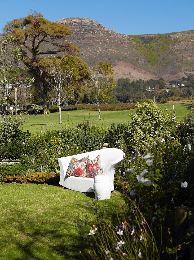 White, outdoor sofa with ethnic scatter cushions in sunny garden in front of mountain landscape