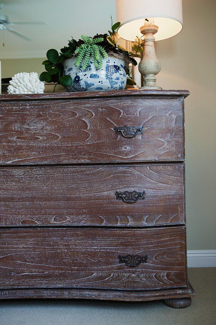 Detail of wooden chest of drawers, house plant and lit lamp against wall; California; USA