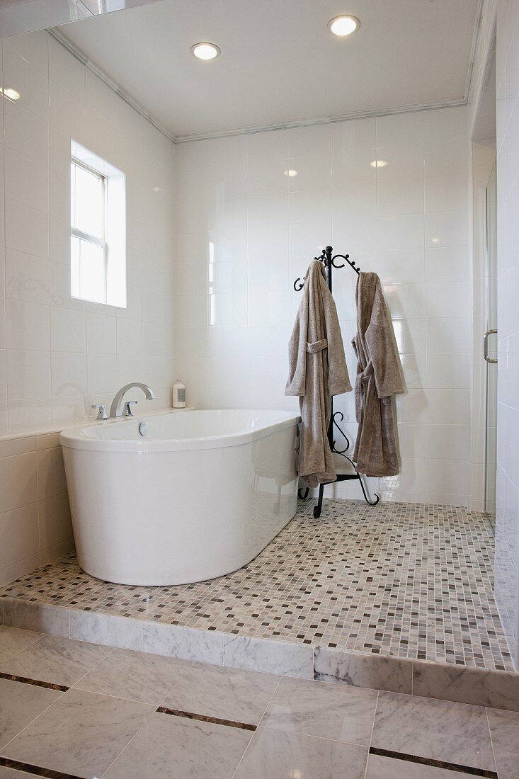 Bademäntel auf Garderobenständer neben gerundeter Badewanne auf Podest mit Mosaikfliesen Boden in weißem Bad mit Lichtreflexionen