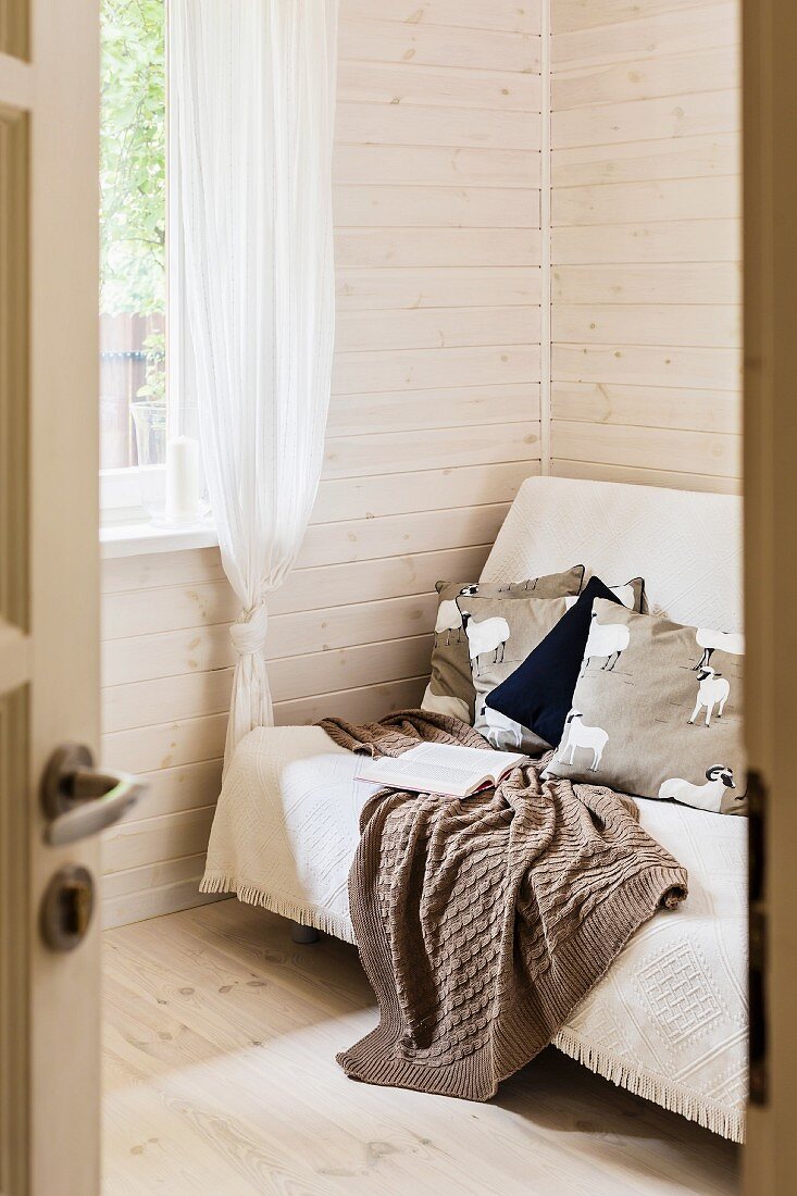 Blick durch offene Tür auf Sofa mit Tagesdecke, in Zimmerecke vor hell lasierter Holzwand