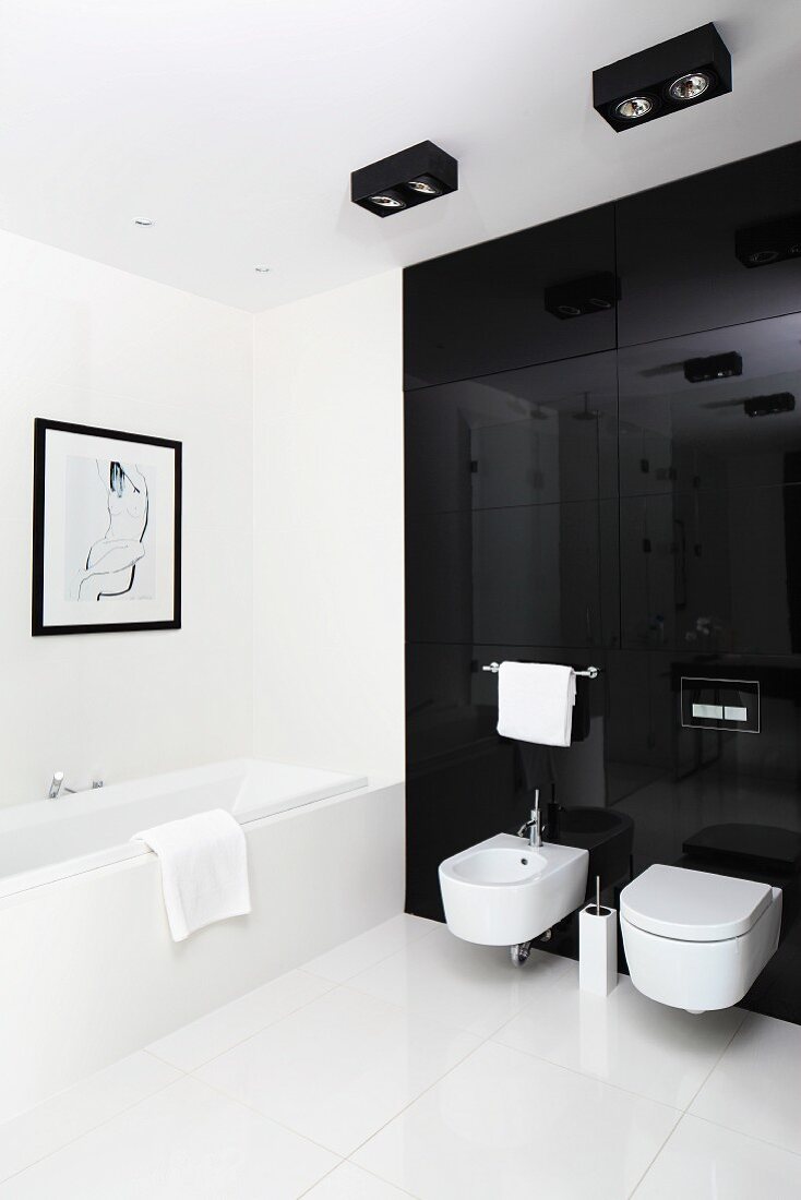 Toilet and bidet mounted on black glass wall and large, white floor tiles in designer bathroom