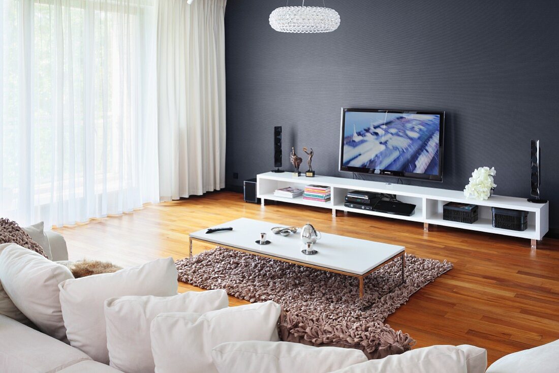 Kissenreihe auf Sofa und Couchtisch auf flokatiartigem Teppichläufer, an grauer Wand weisses Lowboard mit TV, neben Fenster mit luftigem Vorhang
