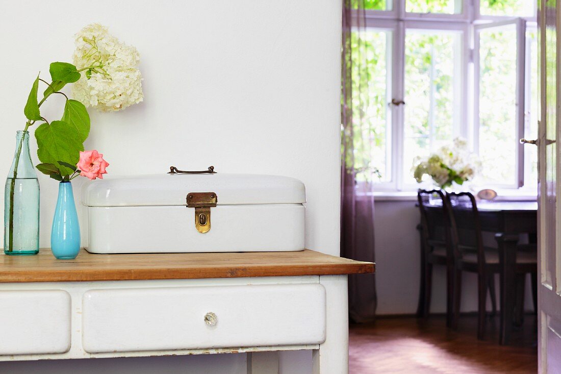 Alter Brotkasten und Blumenvasen auf Wandtisch mit weissen Schubladen, an der Seite offene Tür mit Blick ins Esszimmer auf Tisch am Fenster