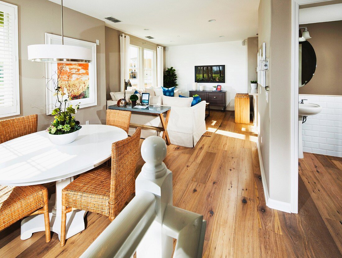 Ovaler, weisser Esstisch und Rattan Stühle auf Holzdielenboden im Hintergrund Loungebereich in offenem Haus