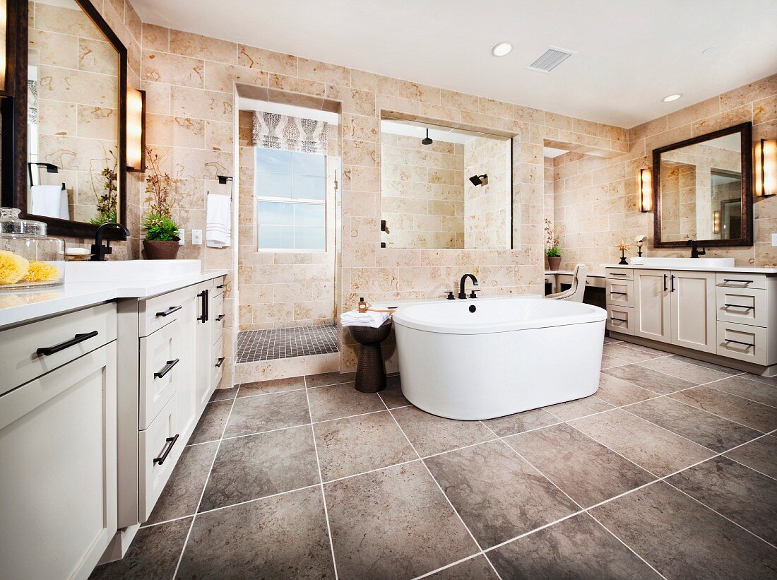 Freistehende Badewanne auf grauen, grossformatigen Fliesen, vor abgetrenntem Duschbereich an den gegenüberliegenden Wandseiten jeweils ein Waschtisch und gerahmter Spiegel