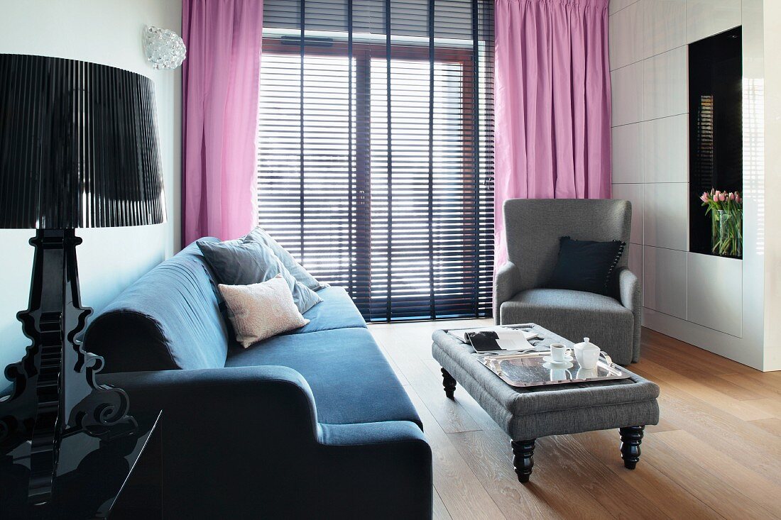 A velvet sofa and an elegant upholstered table in front of floor-length windows with lilac-coloured curtains and half-opened blinds