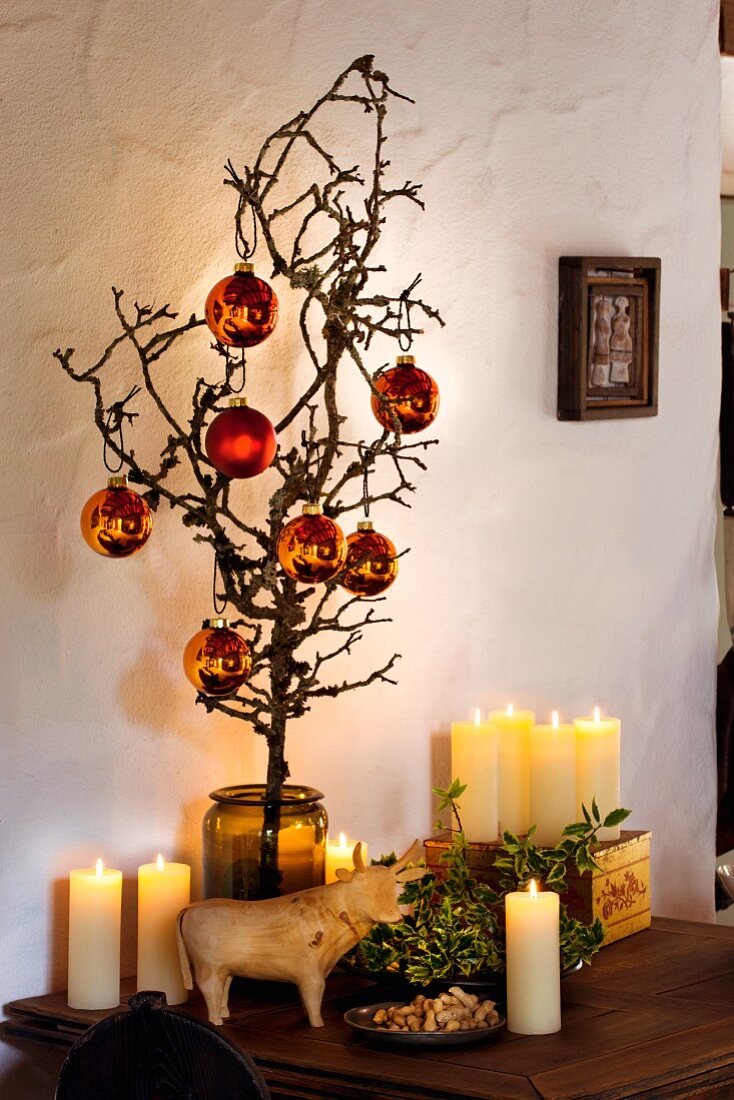 Gnarled fruit tree branch with Christmas tree baubles in glass vase; lit candles and wooden cow
