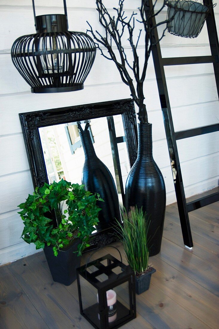 Schwarze Laternen, Spiegel mit schwarz lackiertem Rahmen und schwarze Vase vor weisser Holzwand