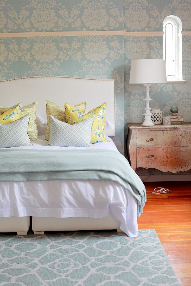 Floral, pastel blue wallpaper and rug combined with timeless mixture of patterns on scatter cushions on double bed