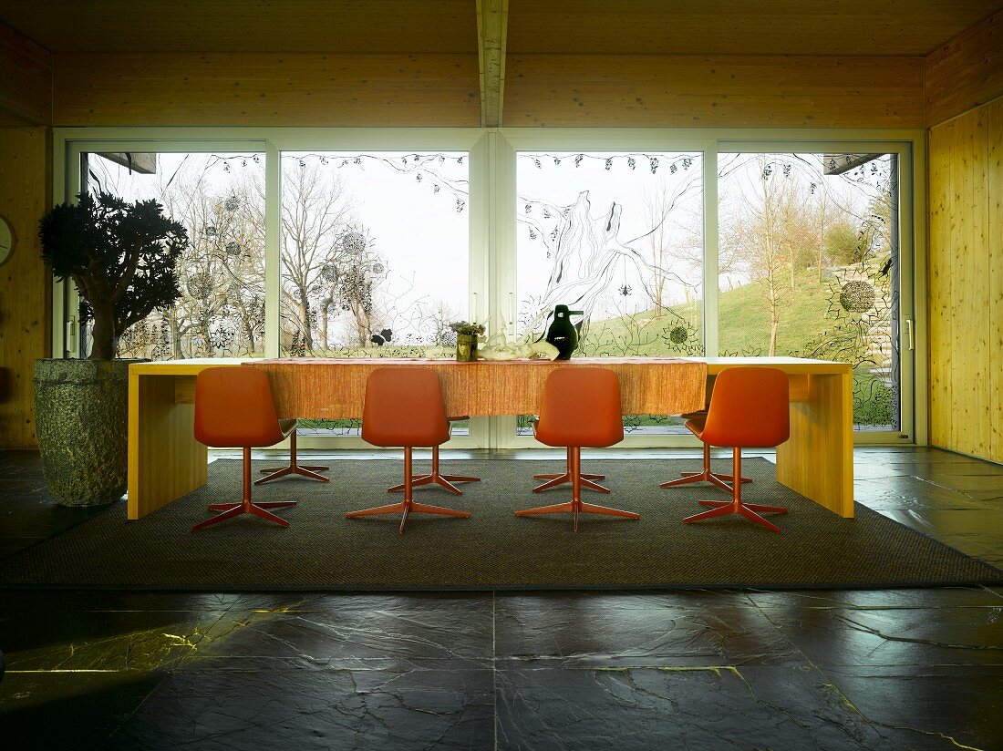Dining room in wooden house with orange shell chairs at long dining table in front of glass wall with view of garden