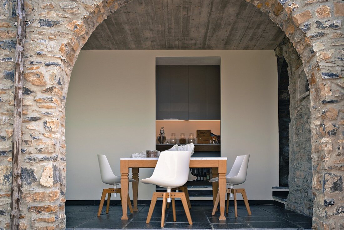 Blick durch die rahmenlose Verglasung eines Natursteinbogens auf Esstisch mit modernen Schalenstühlen; Blick in Küche im Hintergrund