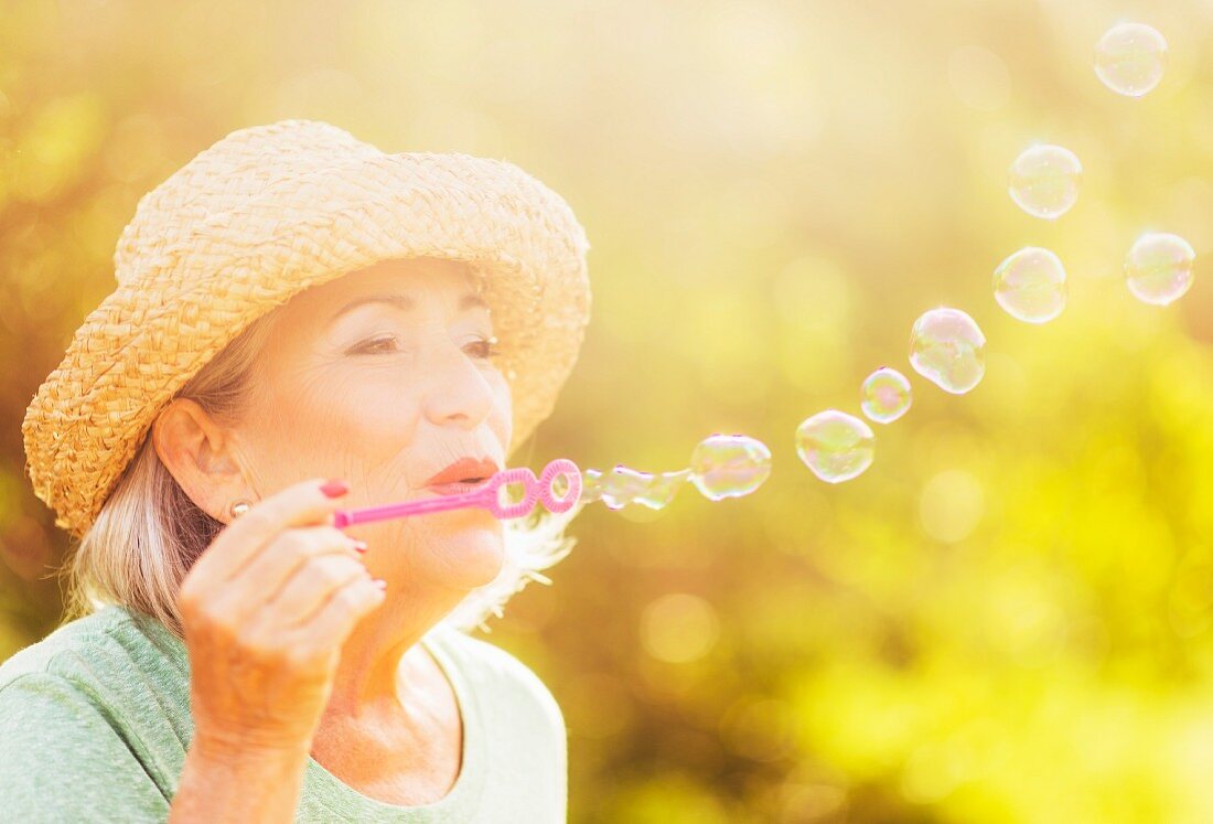 Ältere Frau mit Strohhut bläst Seifenblasen ins spätsommerliche Sonnenlicht