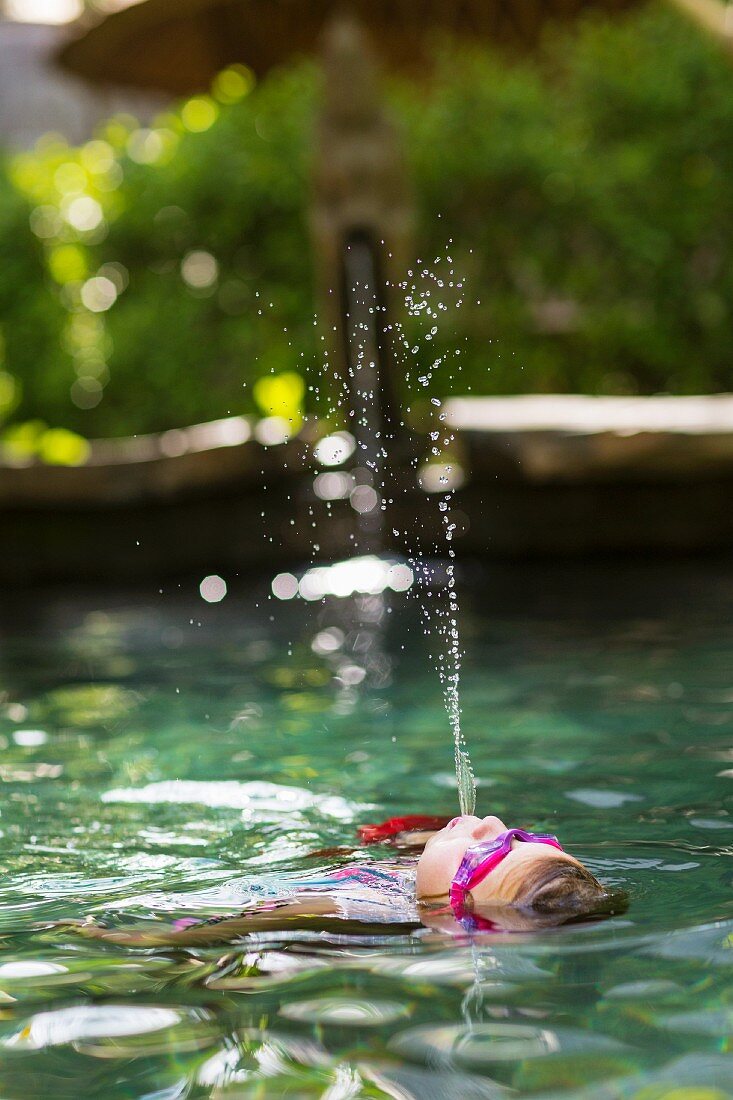 Mädchen mit Schwimmbrille prustet rücklings im Pool liegend Wasser in die Luft