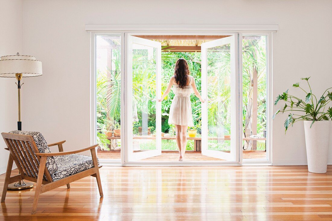Blick von Wohnraum auf junge Frau beim Öffnen der Fenstertüren auf eine Terrasse