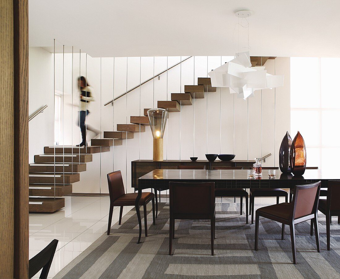 Dining table, upholstered chairs and patterned rug; woman walking down winding staircase with vertical metal balusters