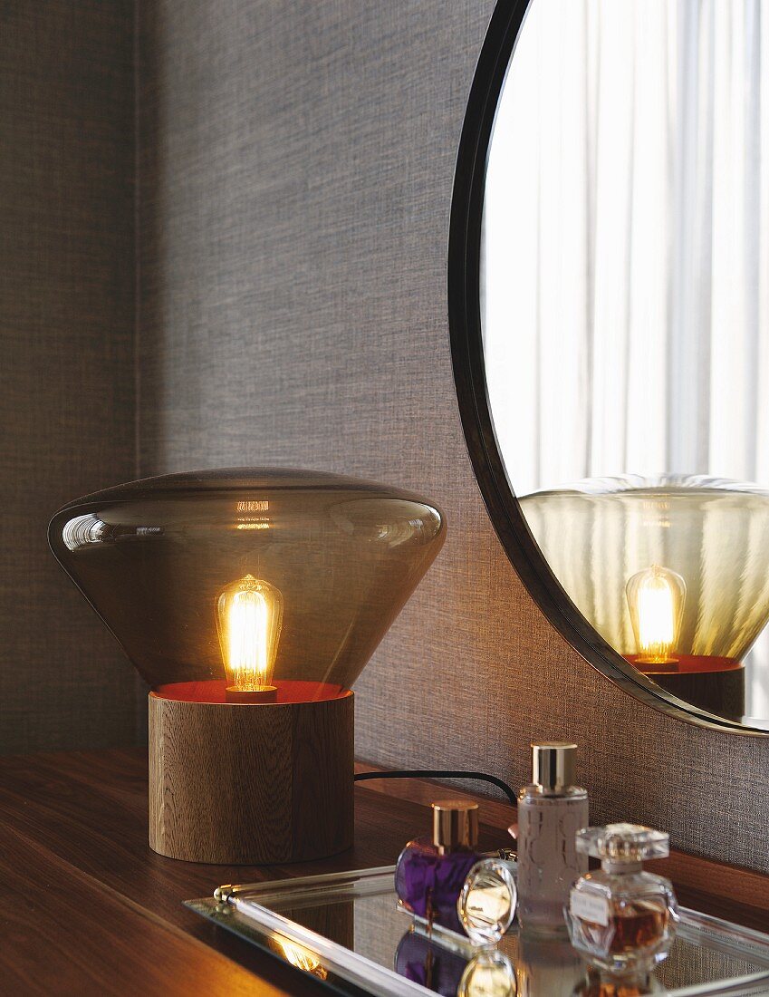 Tischleuchte mit Glasschirm und Parfumflakon auf Tablett, vor Wand mit rundem Spiegel