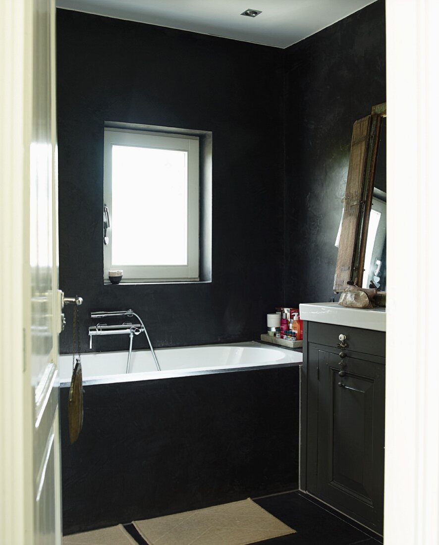 Blick durch offene Tür in schwarz getöntes Bad, seitlich teilweise sichtbarer Waschtisch neben Badewanne unter Fenster