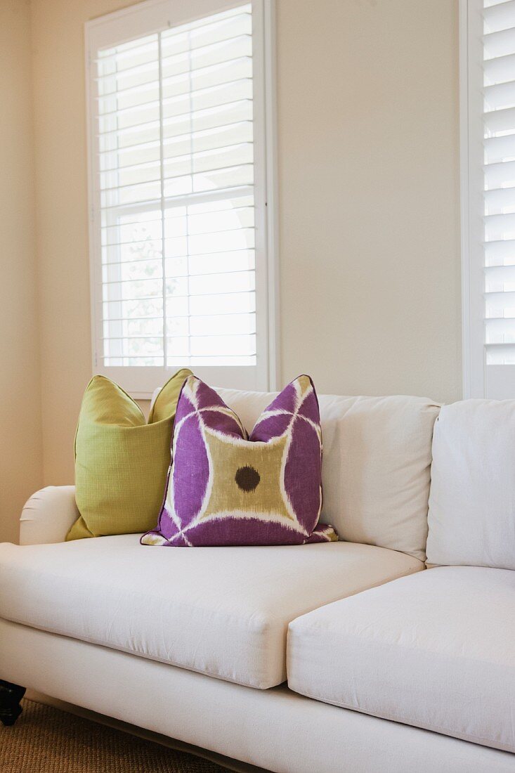 weiße Couch mit lila-grünen Zierkissen vor Lamellenfenstern
