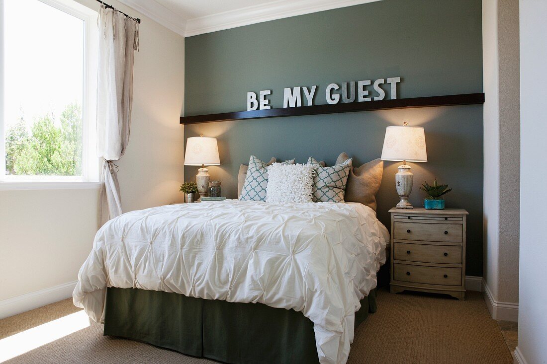 Modernes Gästezimmer mit Doppelbett mit Schriftzug Be my Guest an der grauer Zimmerwand