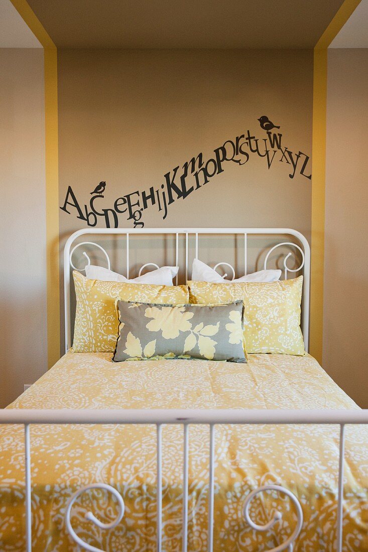 Schlafzimmer mit aufgemaltem Alphabet als Wanddekoration über einem weißem Metallbett mit gelbgemusterter Bettwäsche