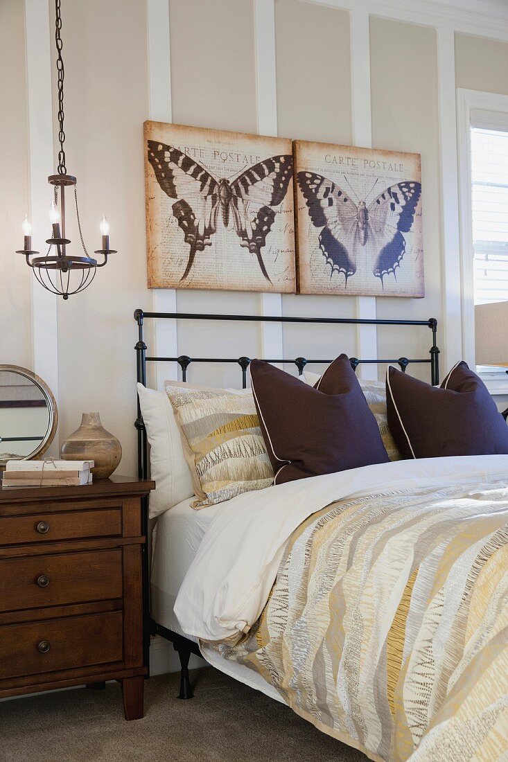Zeitgenössisches Schlafzimmer mit Metallbett, Nachttisch, kleinem Kronleuchter als Nachtischleuchte & Wandbildern mit Schmetterlingsmotiven