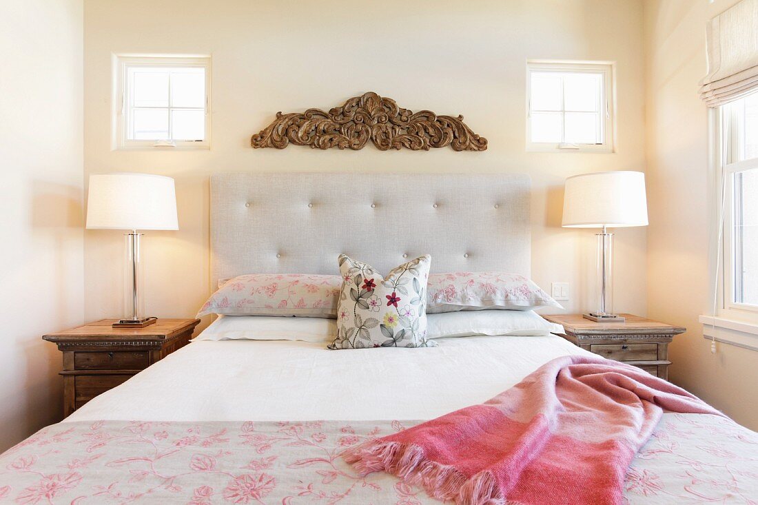 Symmetrisch angeordnetes Schlafzimmer mit Bett eingerahmt von Nachttischen, Tischleuchten, Holzornament & kleinen Fenstern
