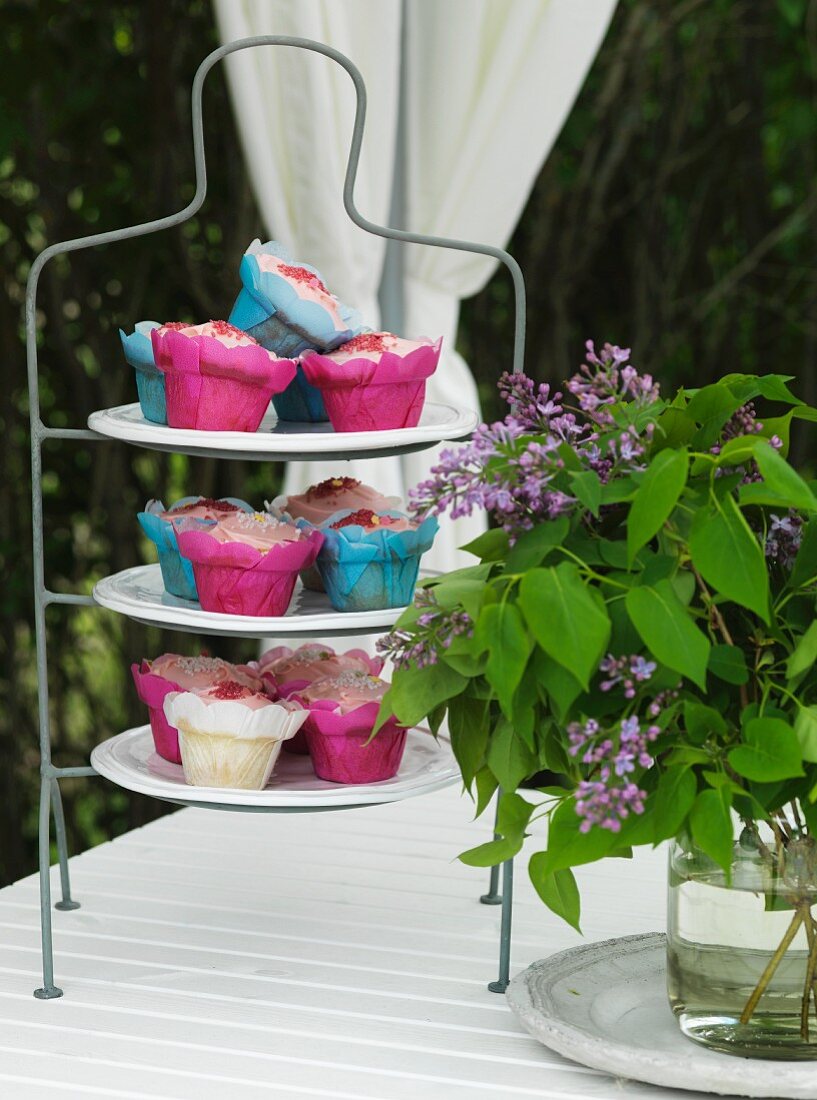 Muffins in colourful paper cases on cake stand and vase of lilac on garden table