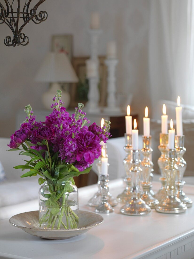 Vase of purple garden flowers and lit candles in silver candlesticks on table