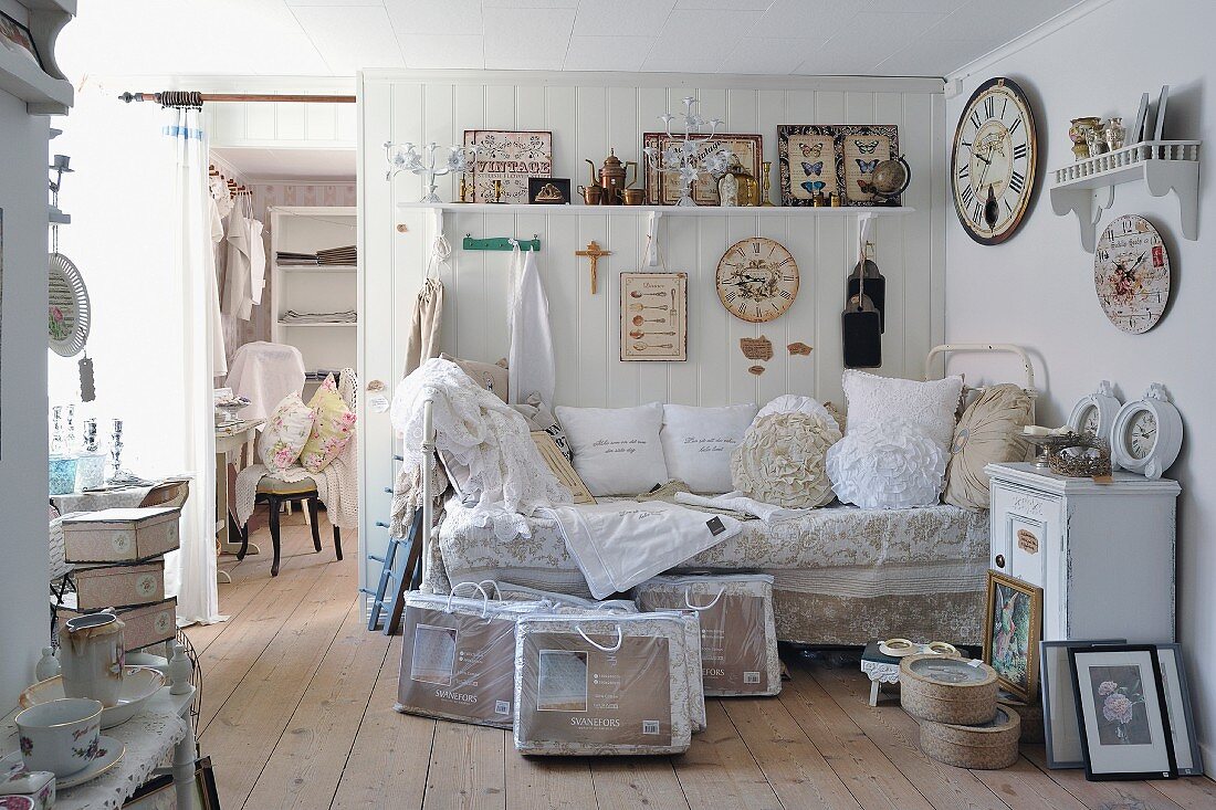 Tagesbett mit Spitzenkissen vor weisser Holzwand mit Vintage Uhrensammlung und nostalgischen Accessoires auf Konsolenregalen