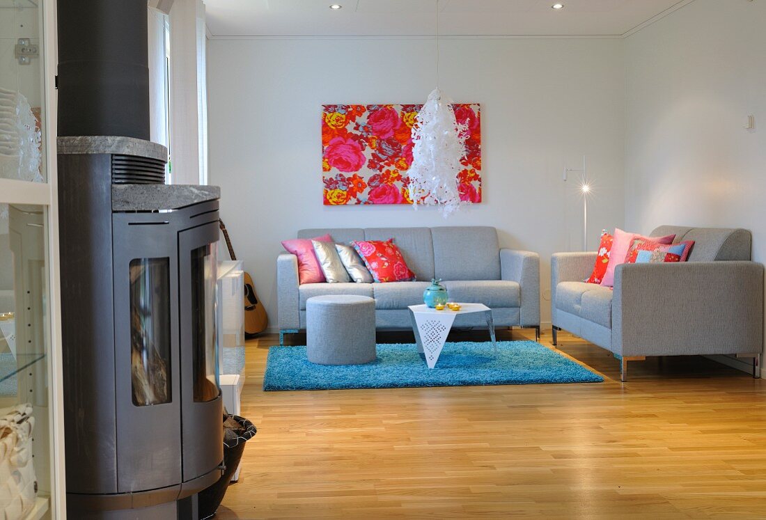 Pale grey, designer sofa set, pale blue rug, modern artwork in shades of red and wood-burning stove in foreground