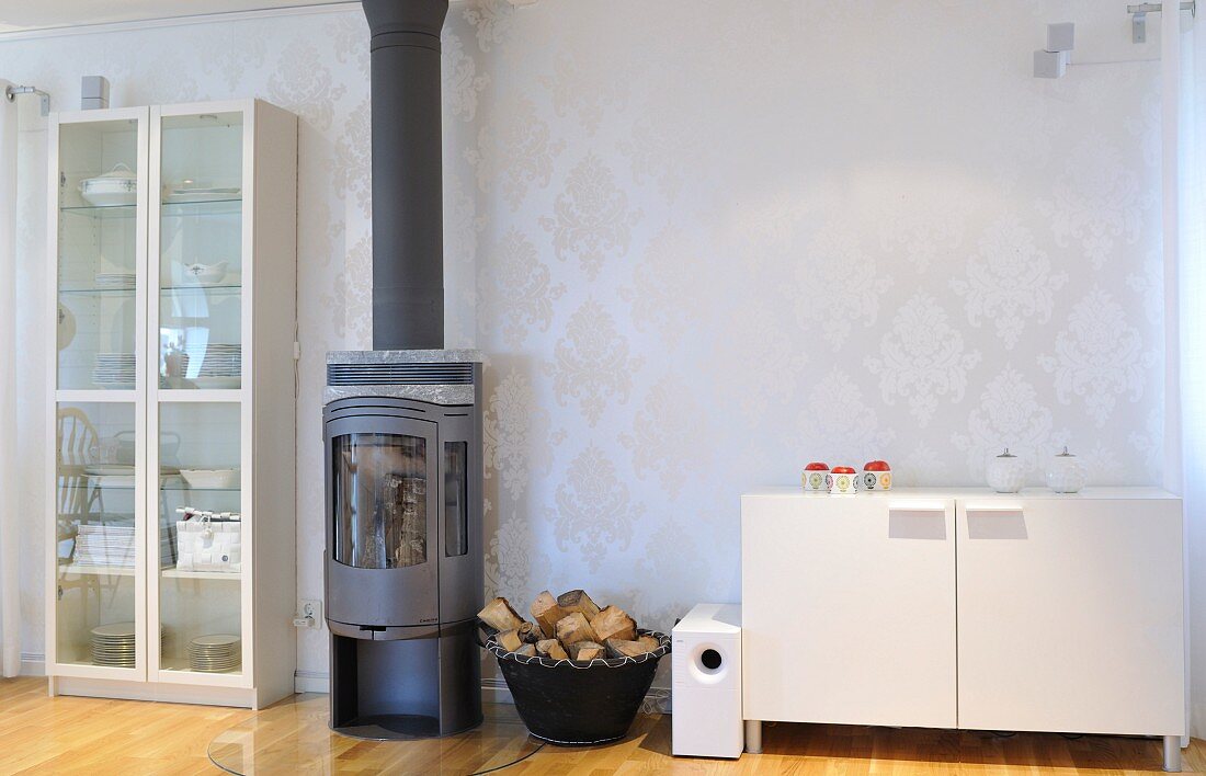 Wood-burning stove between glass-fronted stove and white sideboard against wall covered in pale brocade wallpaper