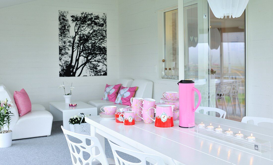 Pinkfarbenes Geschirr, Thermoskanne und Teelichter auf weißem Tisch, im Hintergrund gemütliche Loungeecke